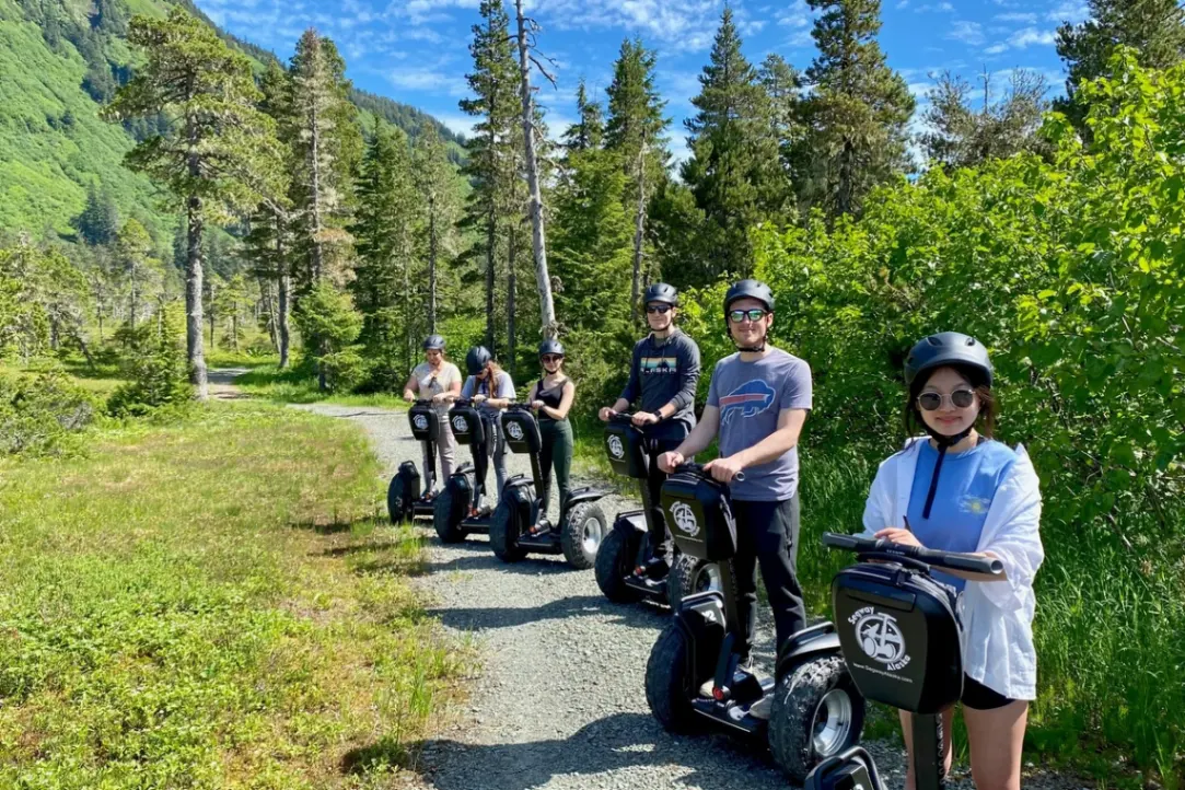 Group on Segways waving by an Alaskan lake with snow-capped mountains in the background.