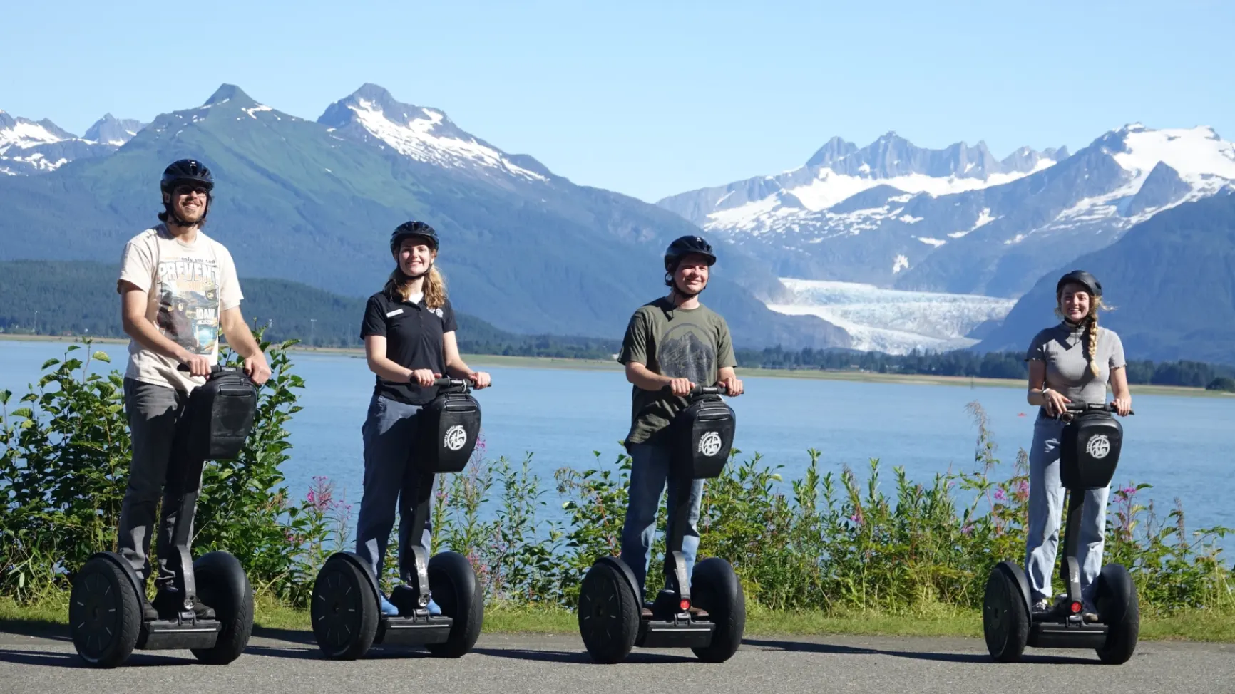 V1-0001_iRide-Website-1920x1080 Two people on Segways waving by the water with snowy mountains in the background during an iRide Alaska tour.